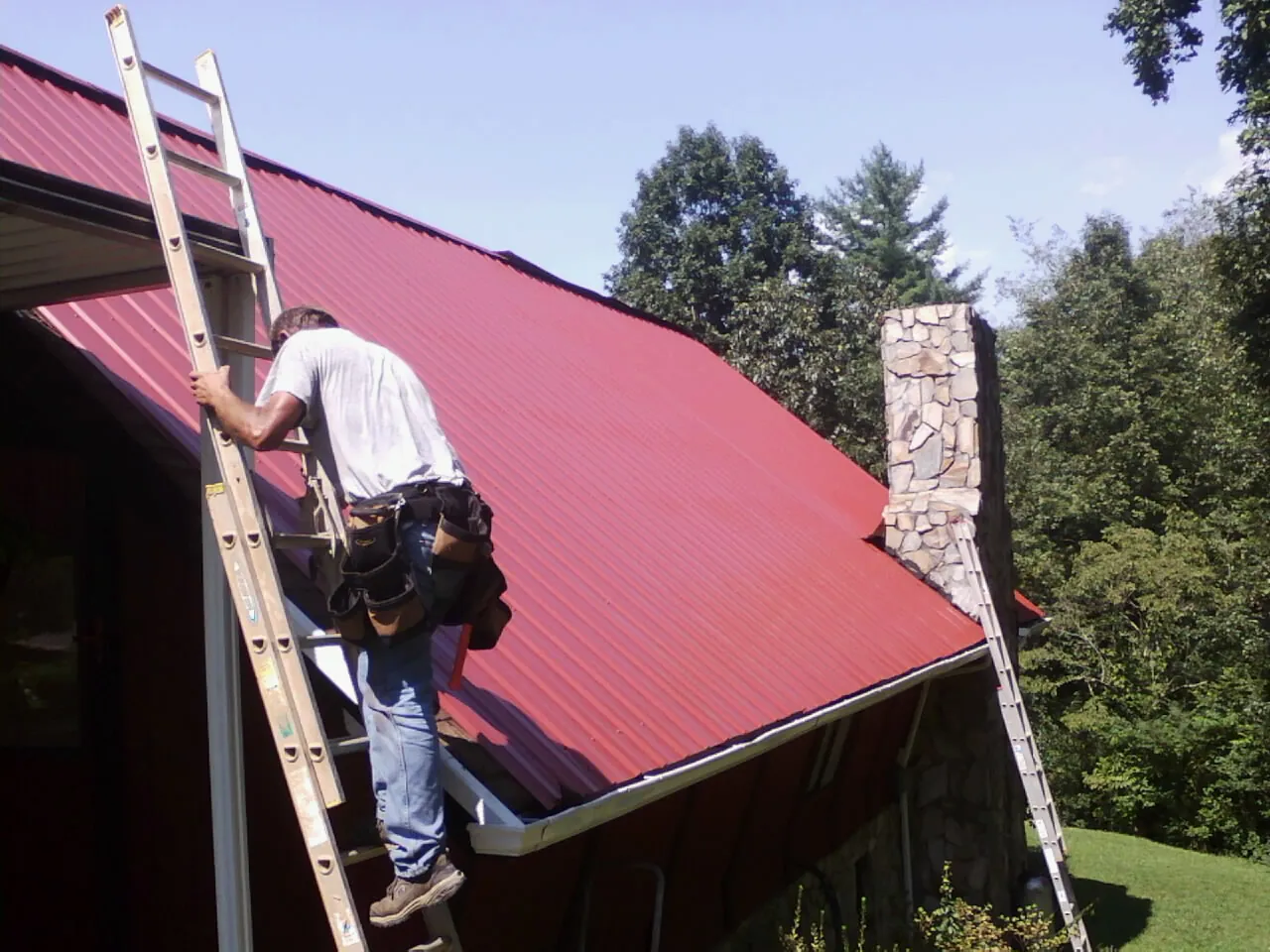 Roofer on a ladder beside a red metal roof and stone chimney — Soffit & Fascia Repair by Roofing Brothers in Artesia